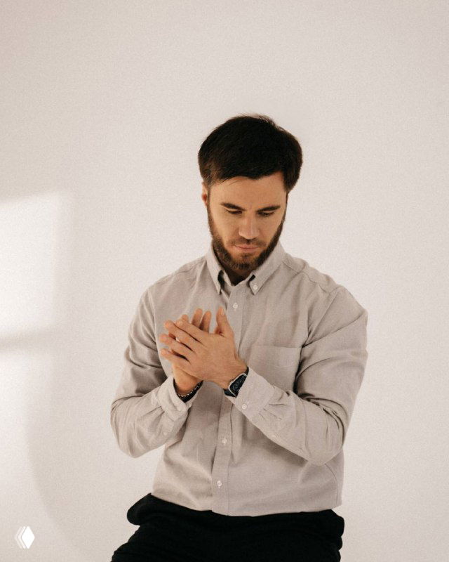 Мужчина в светлой рубашке в нейтральной студии, задумчиво сжимающий руки — иллюстрация делового размышления о контенте и продажах в канале.