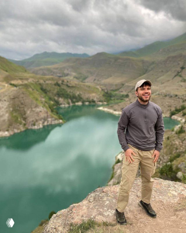 Человек стоит на скалистой кромке над бирюзовым озером и горами, походная сцена с кепкой и ветреной погодой, иллюстрация к посту о маркетинге.