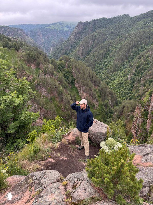 Человек в тёмной куртке и кепке стоит на скалистом выступе, вокруг зелёные склоны и глубокое ущелье Кавказских гор — панорамный пейзаж.