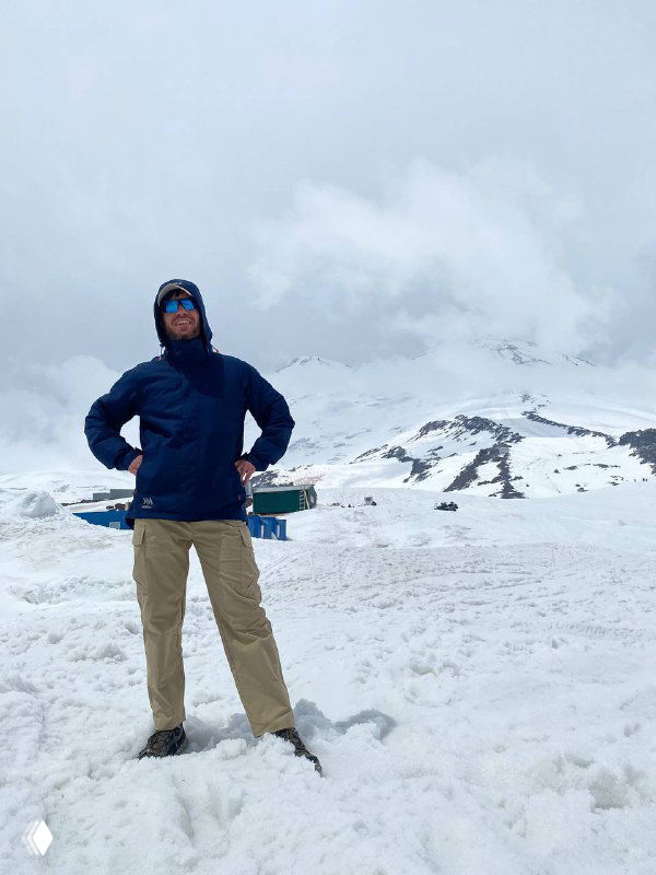 Человек в тёплой куртке стоит на заснеженном склоне, вокруг снежное поле и горные вершины — кадр с высокогорного маршрута и ощущением высоты.