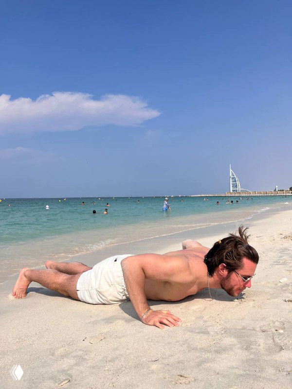 Мужчина на песчаном пляже делает отжимание; на заднем плане виден горизонт с моря и узнаваемый отель Burj Al Arab, солнечная день.
