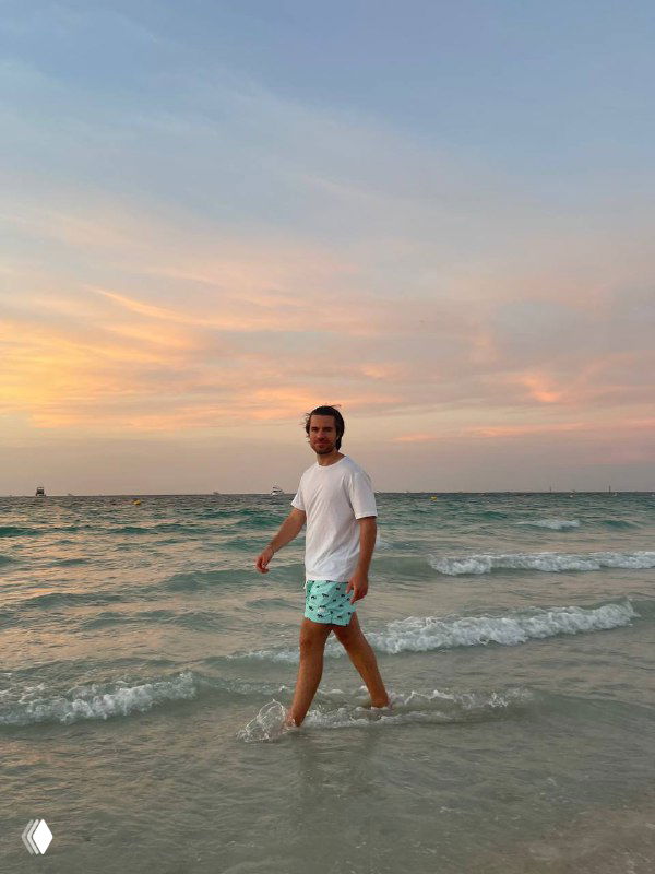 Мужчина в белой футболке и шортах идет по мелководью на закате; виден берег, волны и нежное пастельное небо.