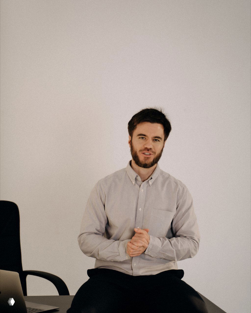 Мужчина в светлой рубашке сидит на столе рядом с ноутбуком и смотрит в камеру; рабочий портрет для темы контент-воронок.