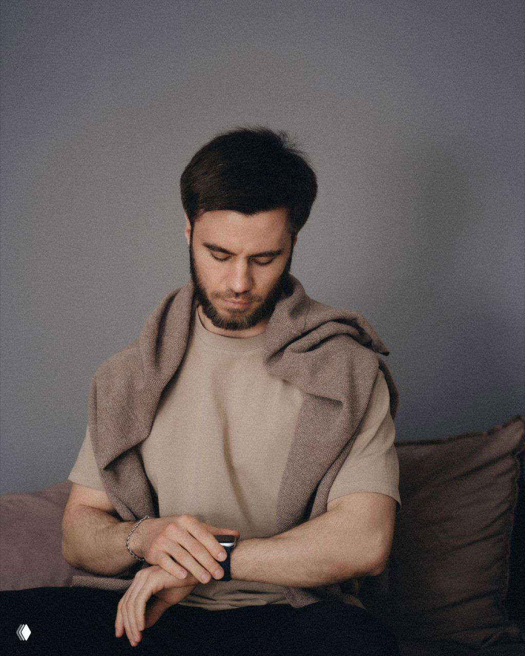 Мужчина в повседневной одежде сидит на диване и смотрит на наручные часы — спокойная фотография для иллюстрации мысли о дисциплине и постепенном росте в рекламе.