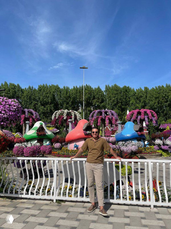 Мужчина фотографируется у белой ограды на фоне большой цветочной инсталляции и зелёных деревьев в солнечный день в парке