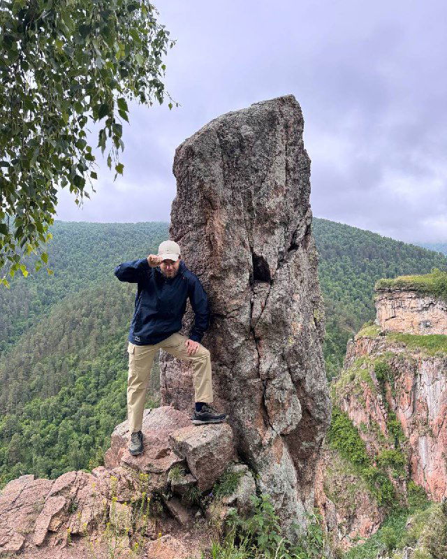 Человек в кепке и куртке стоит на краю скалы на фоне зелёных гор и облачного неба, символизируя подготовку к сезону продаж.
