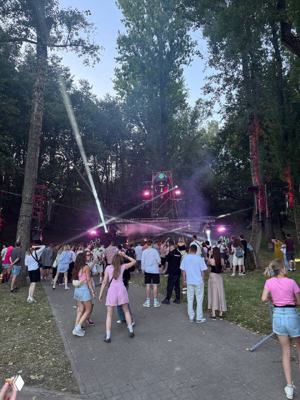 Фотография лесной сцены музыкального фестиваля: диджейская платформа с яркими прожекторами и дымом, люди танцуют и собираются вдоль дорожки у сцены.