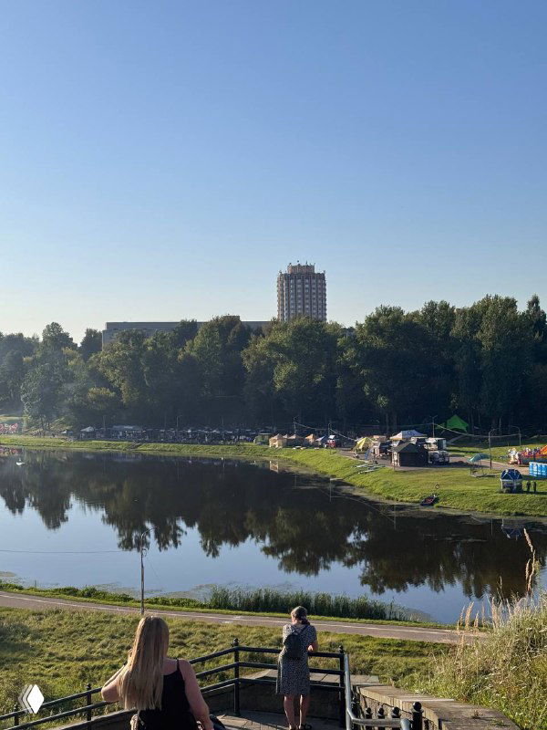 Панорама береговой зоны фестиваля у озера: отдыхующие на траве и берегу, палатки и сцены на фоне зелёных деревьев и высотного дома вдали, тихий летний пейзаж.