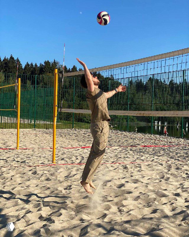 Игрок в хаки прыгает у сетки на песчаной площадке, бьёт мяч для пляжного волейбола; на заднем плане лес и голубое небо.
