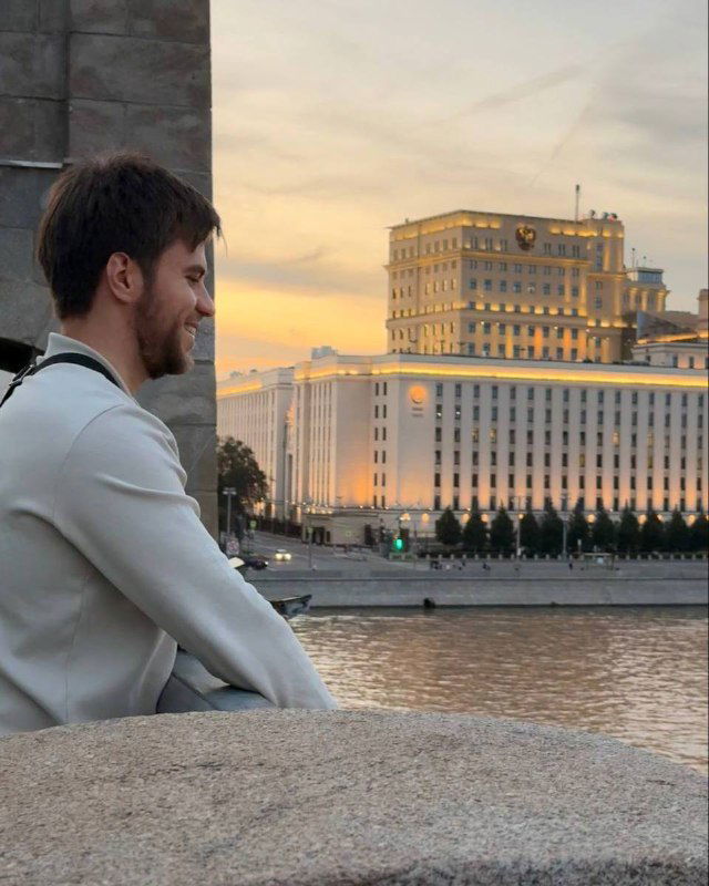 Человек сидит на набережной у реки в сумерках, на заднем плане видно большой административный дом и городской пейзаж на закате.