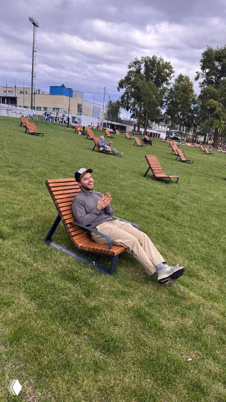 Человек отдыхает на лежаке в городском парке: зелёный газон, несколько шезлонгов, деревья и облачное небо — кадр после рабочего цикла на свежем воздухе.