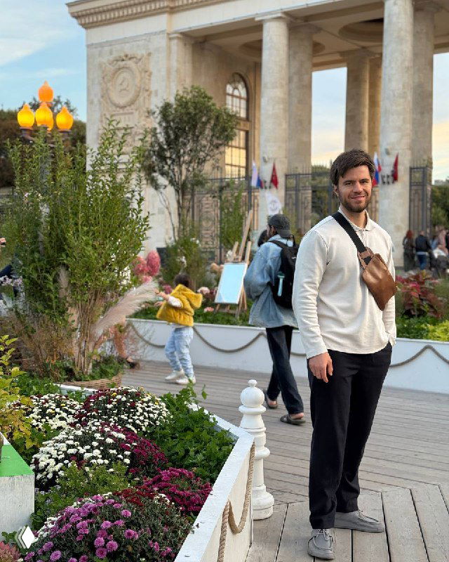 Мужчина в светлой рубашке с поясной сумкой стоит на пешеходной аллее у цветочных клумб, на фоне арки и гуляющих людей.