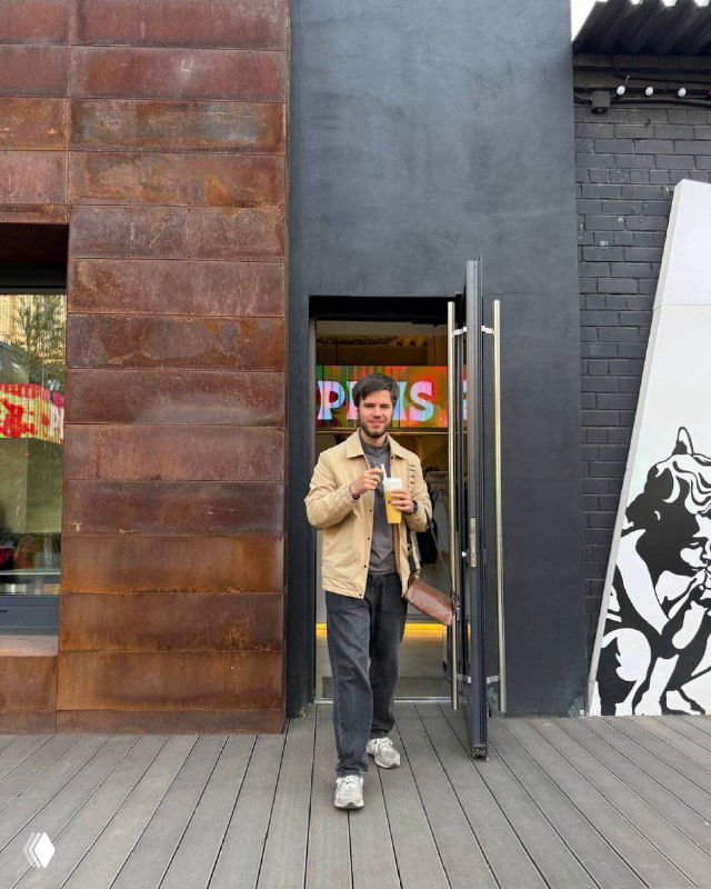 Мужчина в куртке с напитком стоит у входа в заведение на фоне урбанистической стены с граффити — неформальный уличный кадр.