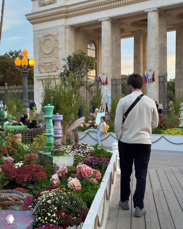 Мужчина в светлой куртке идёт по украшенной цветами аллее рядом с декоративными фонарями; на заднем плане — большая арка.