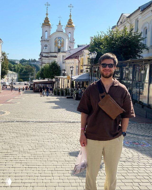 Человек в солнцезащитных очках и сумкой на плече стоит на пешеходной улице с уличными кафе и церковью на заднем плане, солнечный день.