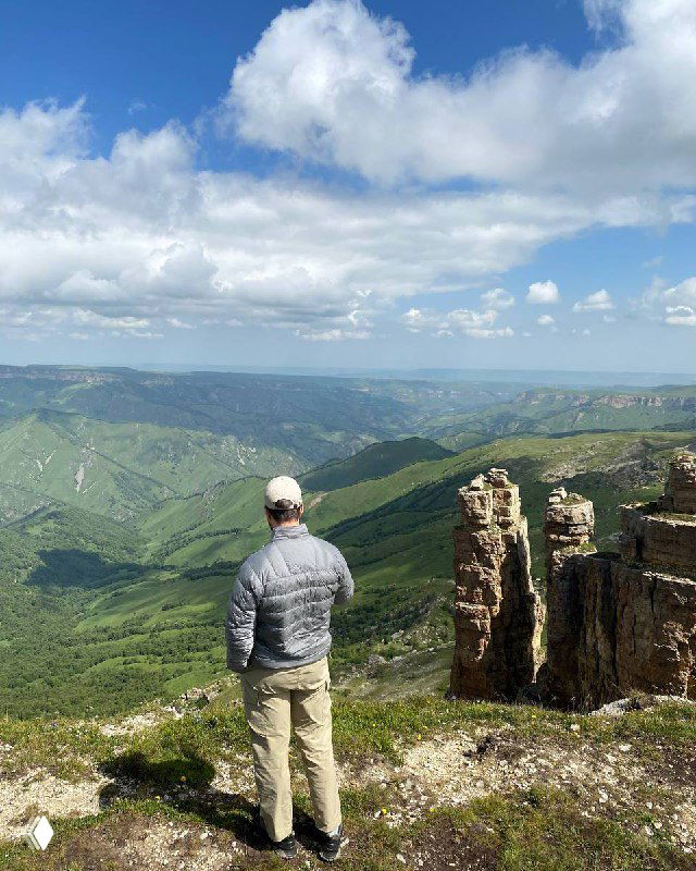 Мужчина в кепке и лёгкой куртке стоит на краю скалы и смотрит на широкий горный ландшафт с долиной под переменным облачным небом.