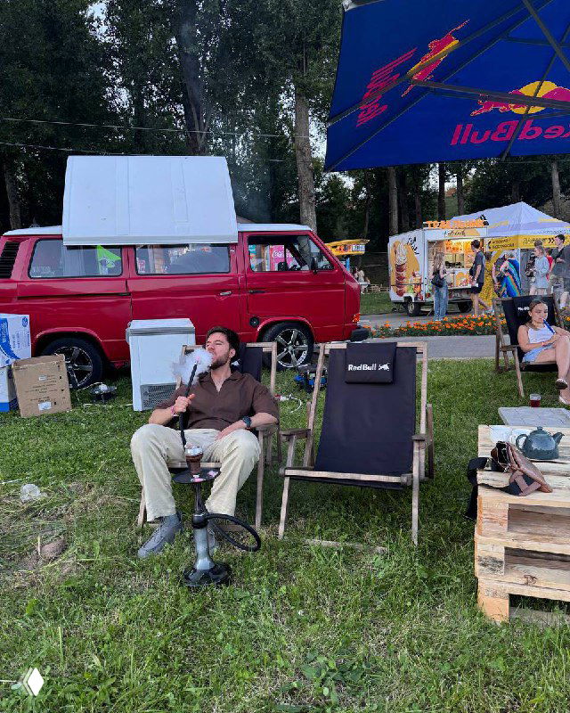 Мужчина отдыхает в раскладном кресле на летнем фестивале у красного фургона, рядом зонтик Red Bull и палатками с едой.