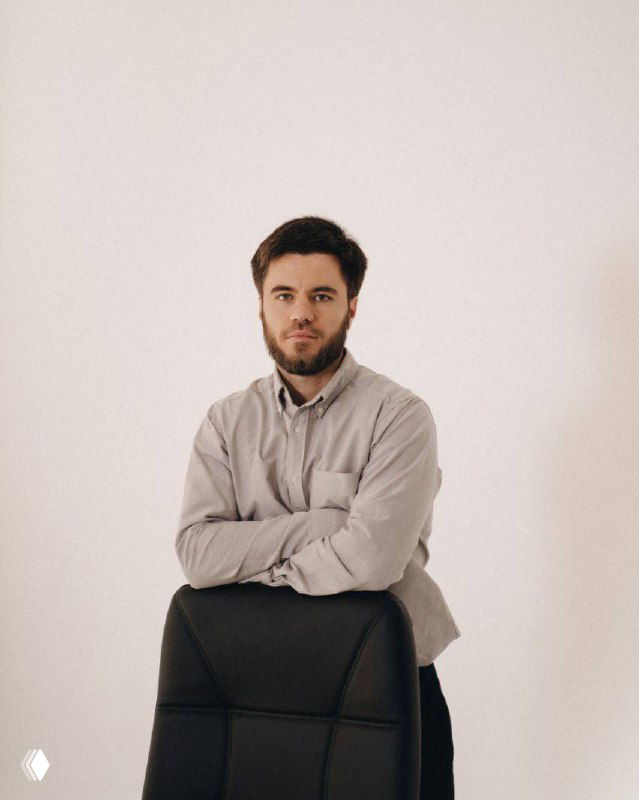 Портрет мужчины в светлой рубашке за креслом на однотонном светлом фоне; нейтральная деловая фотография для иллюстрации материала о рекламе в Telegram.