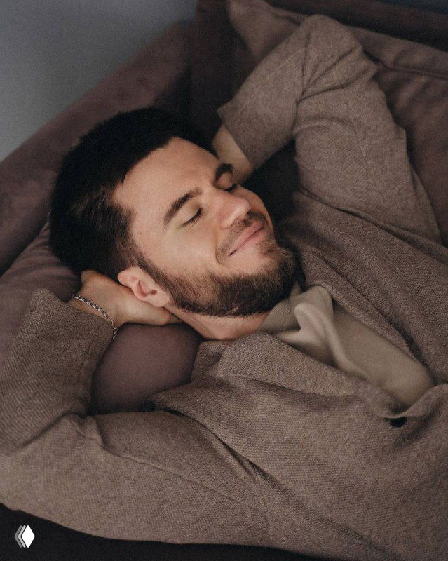 Портрет молодого мужчины в уютном свитере на диване, расслабленная поза и лёгкая улыбка — атмосферное фото для иллюстрации авторского поста.