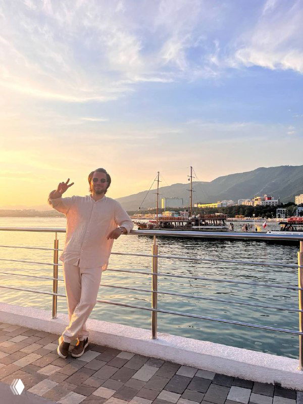 Человек в светлой одежде на набережной у моря на закате, приветственно машет рукой; на заднем плане причал с яхтами и горы, сцена иллюстрирует атмосферу поста о маркетинге