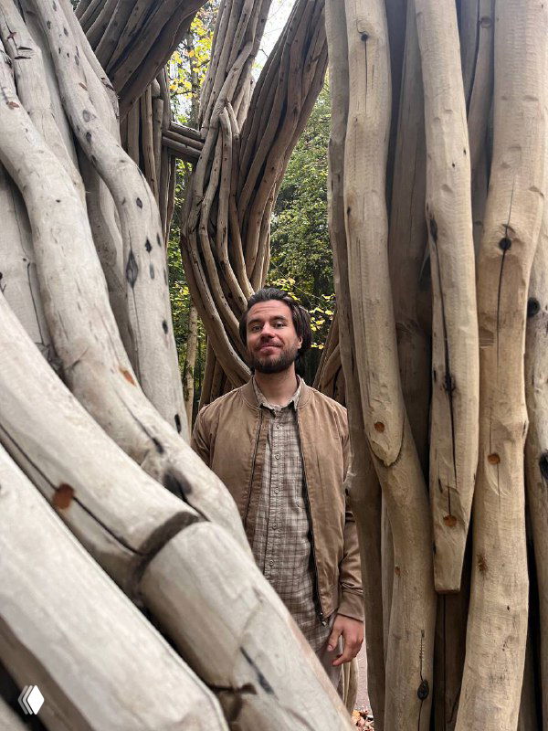 Мужчина стоит между крупными переплетёнными стволами деревьев в парке — портрет на фоне природной композиции с выразительной текстурой древесины.