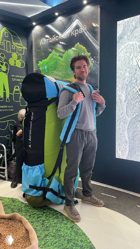 Мужчина в спортивной одежде позирует с крупным туристическим рюкзаком/парапланным снаряжением в выставочном стенде; видны стенды и демонстрационный макет.