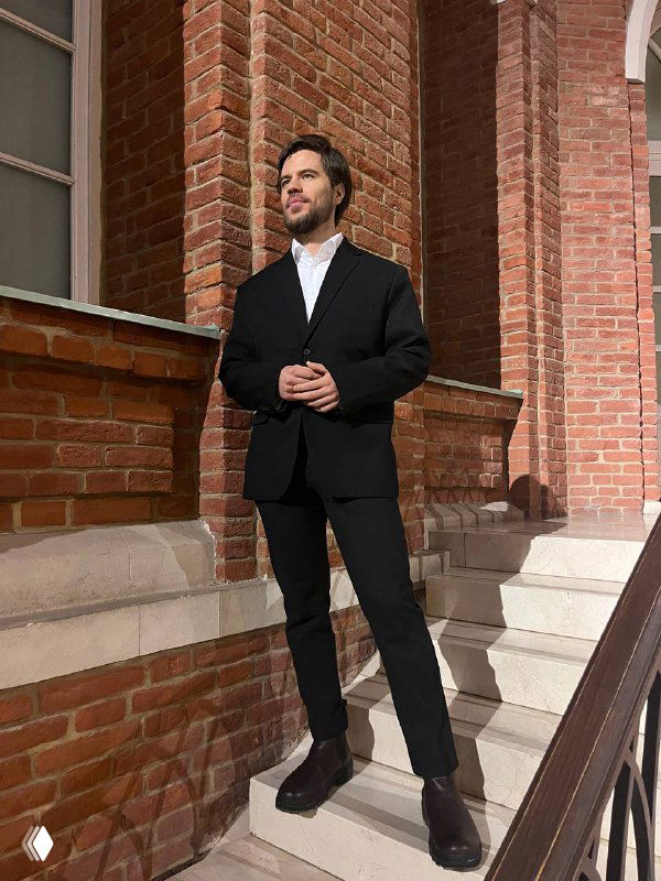 Человек в деловом костюме стоит на ступенях у кирпичного здания; поза и одежда подчёркивают деловой контекст.