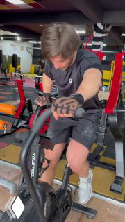 Человек в спортзале на велотренажёре в перчатках; кадр показывает тренировочную обстановку, аксессуары и экипировку, акцент на руках и перчатках.