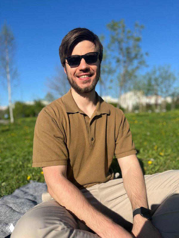 Мужчина в тёмных очках и коричневой рубашке сидит на траве в солнечном парке, улыбается; на фоне деревья и голубое небо.