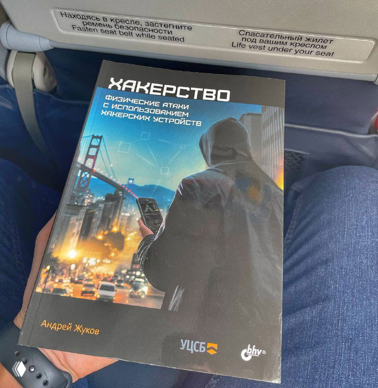 Фотография обложки книги «Хакерство. Физические атаки с использованием хакерских устройств» на коленях у пассажира в самолёте, видна иллюстрация хакера и городской мост.