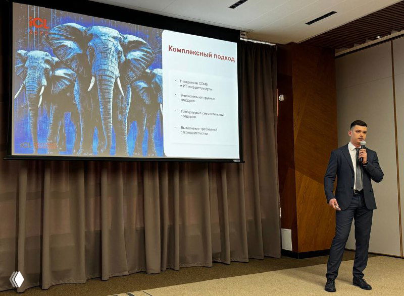 Спикер в деловом костюме на сцене рядом с экраном, на слайде изображение слонов; конференц‑зал и презентация по информационной безопасности.
