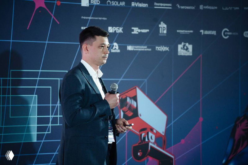 Докладчик у стенда на Positive Hack Days: мужчина в костюме с микрофоном на фоне брендированной графики и логотипов.