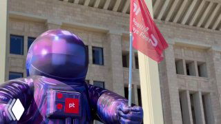 Фигурка талисмана PHDays в фиолетовом скафандре на фоне фасада и флагов фестиваля у входа в Лужники, атмосфера подготовки к событию.