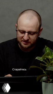 Чёрно‑белый кадр: человек за столом наклонён над планшетом/бумагой, рядом горшок с растением; на изображении видна подпись «Старайтесь».