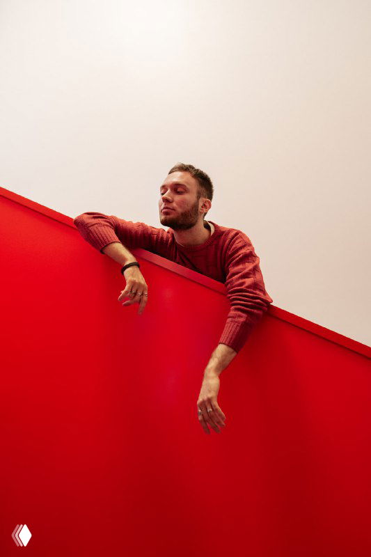 Мужчина в красном свитере опирается на ярко-красную перегородку на нейтральном фоне; портрет в стиле минимализма, представляющий автора канала по инфографике.