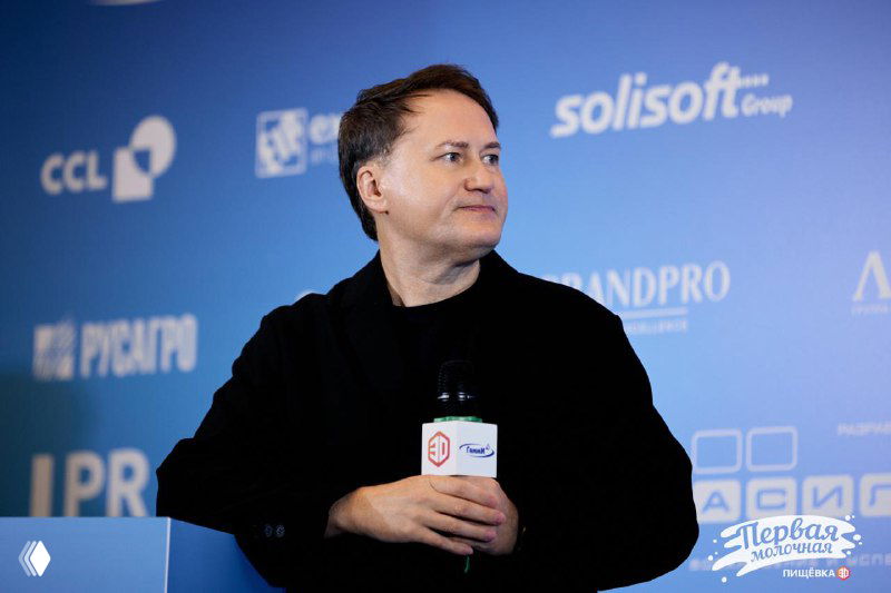 Оратор на сцене конференции на фоне синего баннера с логотипами и названиями, держит микрофон и смотрит в сторону аудитории