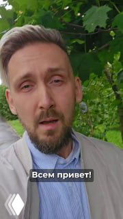 Вертикальное превью: мужчина в пиджаке говорит в камеру на фоне зелёной листвы, внизу виден титр «Всем привет!», атмосферное освещение.