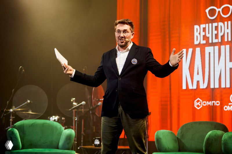 Ведущий на сцене во время шоу «Вечерний Калинин» на Meat Night в Москве: красная сцена, софиты, микрофон и внимательная аудитория за зелёными креслами.