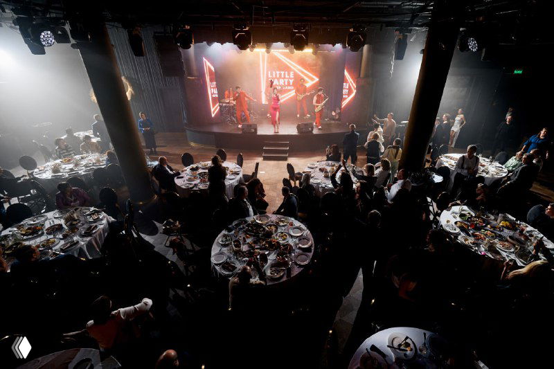 Широкий план банкетного зала с сервированными столами и сценой: атмосфера торжества, гости за столами и сцена с выступающими и световыми эффектами.