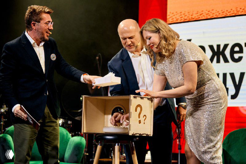 На сцене ведущие и участники вытягивают предметы из коробки в интерактивном номере: шоу-элемент с реквизитом и вовлечённой публикой в зале.