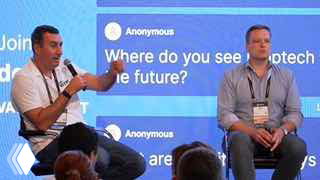 Two speakers on stage at a proptech event, sitting with microphones while a projected slide about proptech appears behind them, audience visible in foreground.