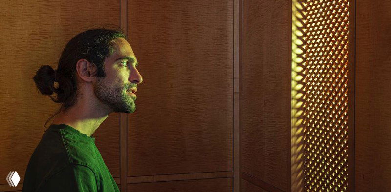 Man in a wood-paneled confessional booth looking toward a lit lattice screen, illustrating the AI-Jesus hologram installation in Switzerland.