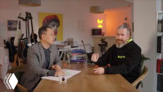 Two men seated at a wooden table in a casual studio, conversing in an interview about AI, UBI and happiness.