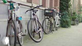 Row of parked city bicycles along a sidewalk near buildings, one with a front basket — illustrative photo for Google’s autonomous/self-driving bicycle news.