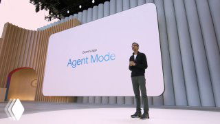 Photograph of a tech conference stage with a presenter beside a large slide that reads 'Agent Mode', illustrating an AI agent demo and product positioning context.