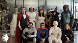 Placy team group photo: employees in Halloween costumes posing in an office with city skyline backdrop, celebrating.