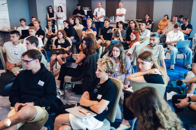 Hackathon audience in a conference room: young founders, students and mentors listening attentively, some taking notes and using laptops during presentations.