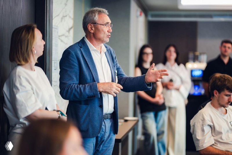 Speaker addressing the room during the hackathon: an investor or mentor presenting to participants with attentive attendees in the background.