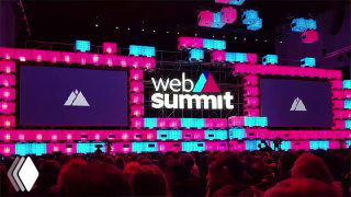 Web Summit stage in pink and cyan lighting showing 'web summit' logo on large screens, audience silhouettes and event atmosphere in Lisbon.
