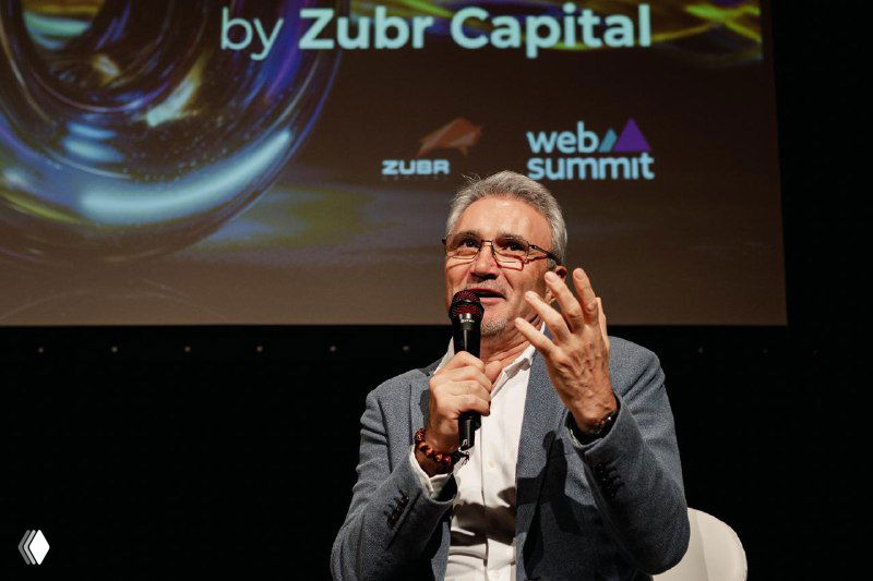 Elderly speaker gesturing while addressing the audience from stage at the Future Unicorns Party 2025, spotlight and event backdrop visible.
